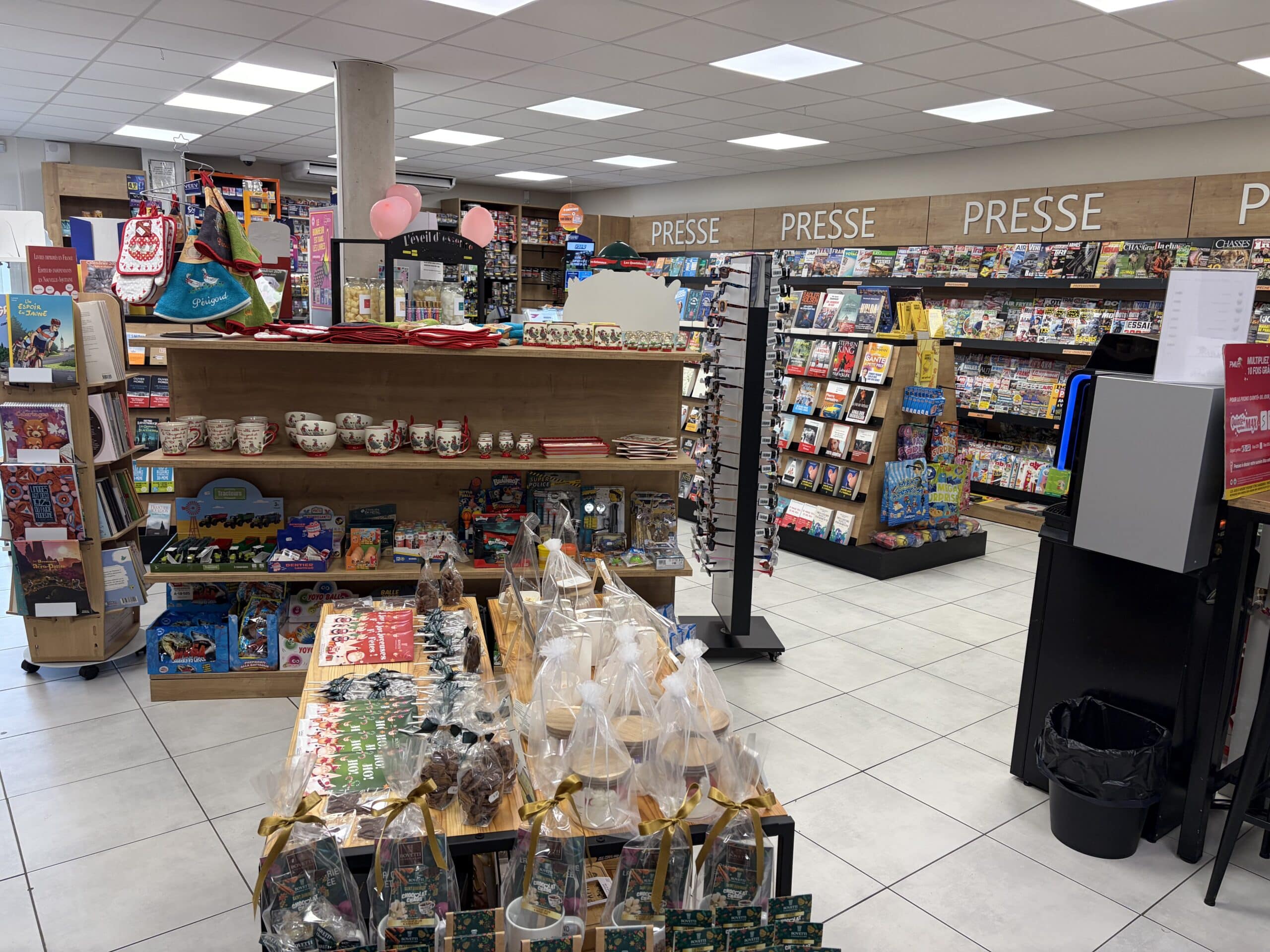 Intérieur d'un magasin de presse et cadeaux avec rayons de magazines, souvenirs du Périgord et confiseries artisanales.