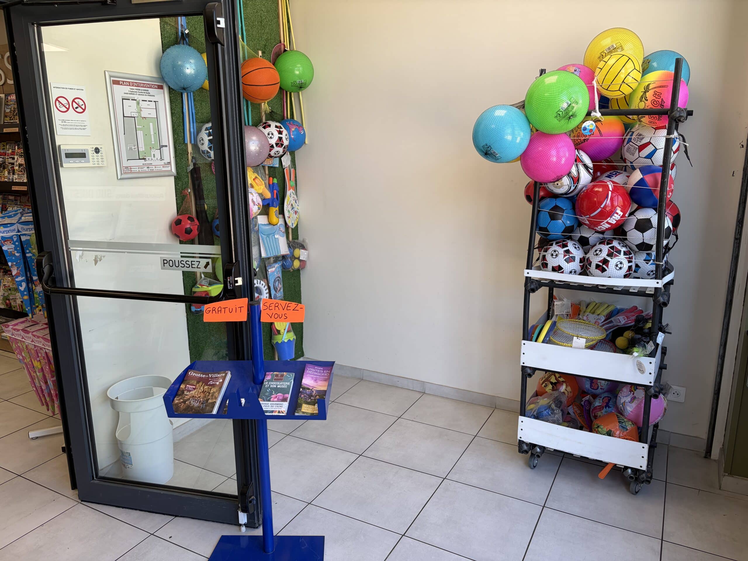 Entrée de magasin : ballons de sport colorés, jouets de plage et brochures gratuites avec panneau Servez-vous.