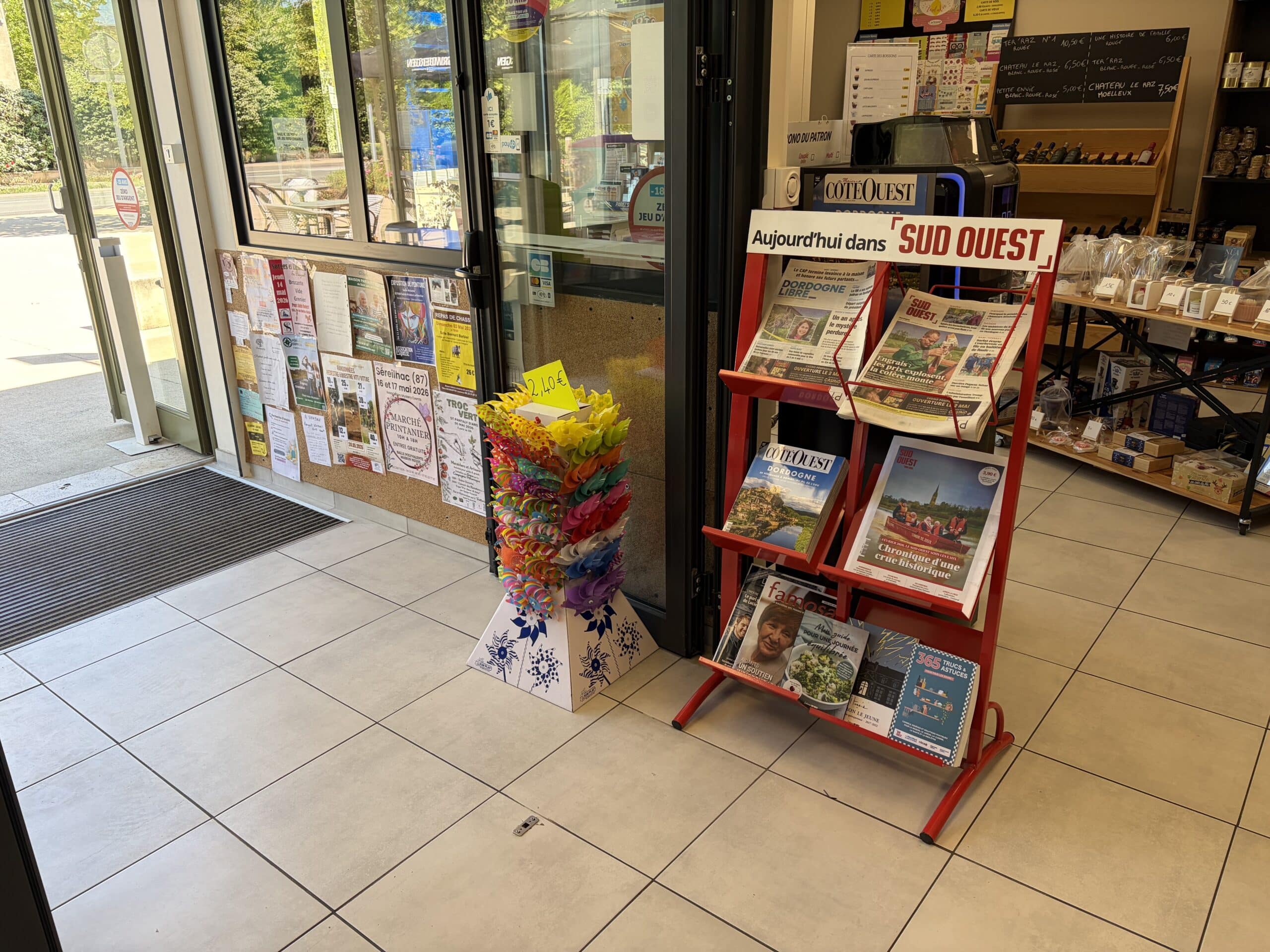 Entrée de boutique avec présentoir Sud Ouest, moulins à vent colorés et mur d'affiches publicitaires.